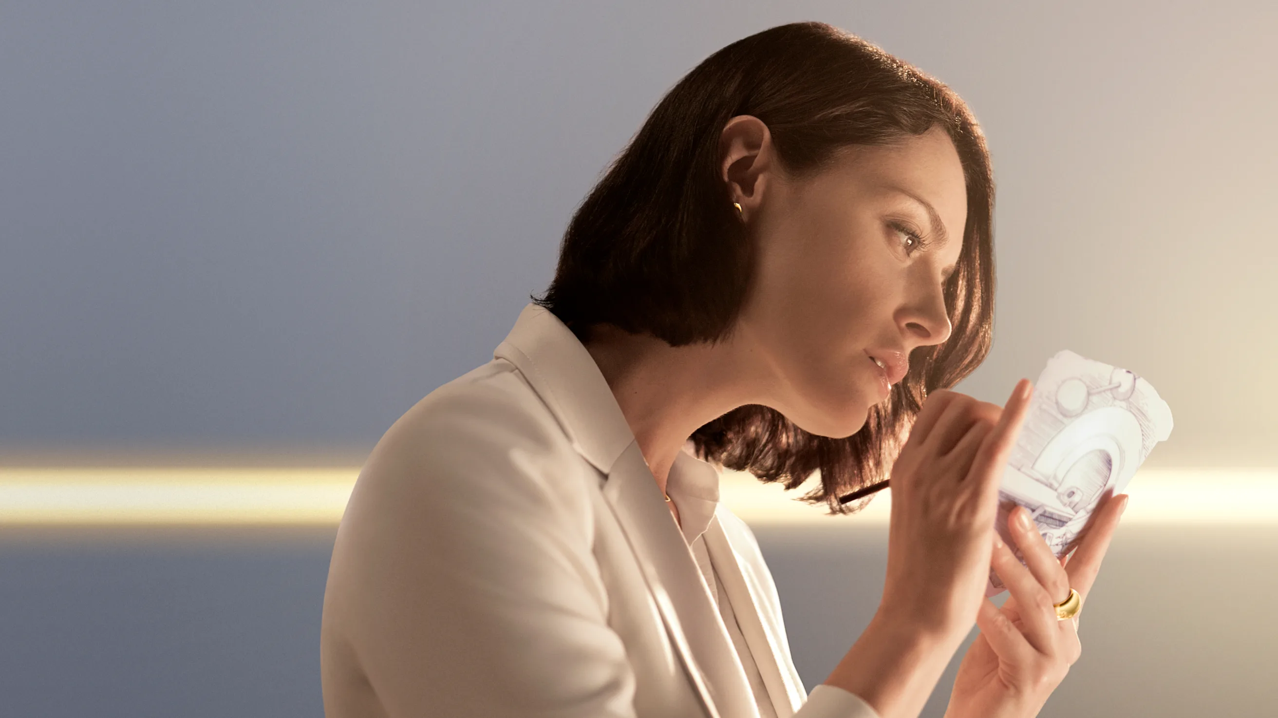 A woman with short brown hair, dressed in a white suit, examines a translucent, futuristic object with architectural designs, set against a softly lit background with a warm glow.