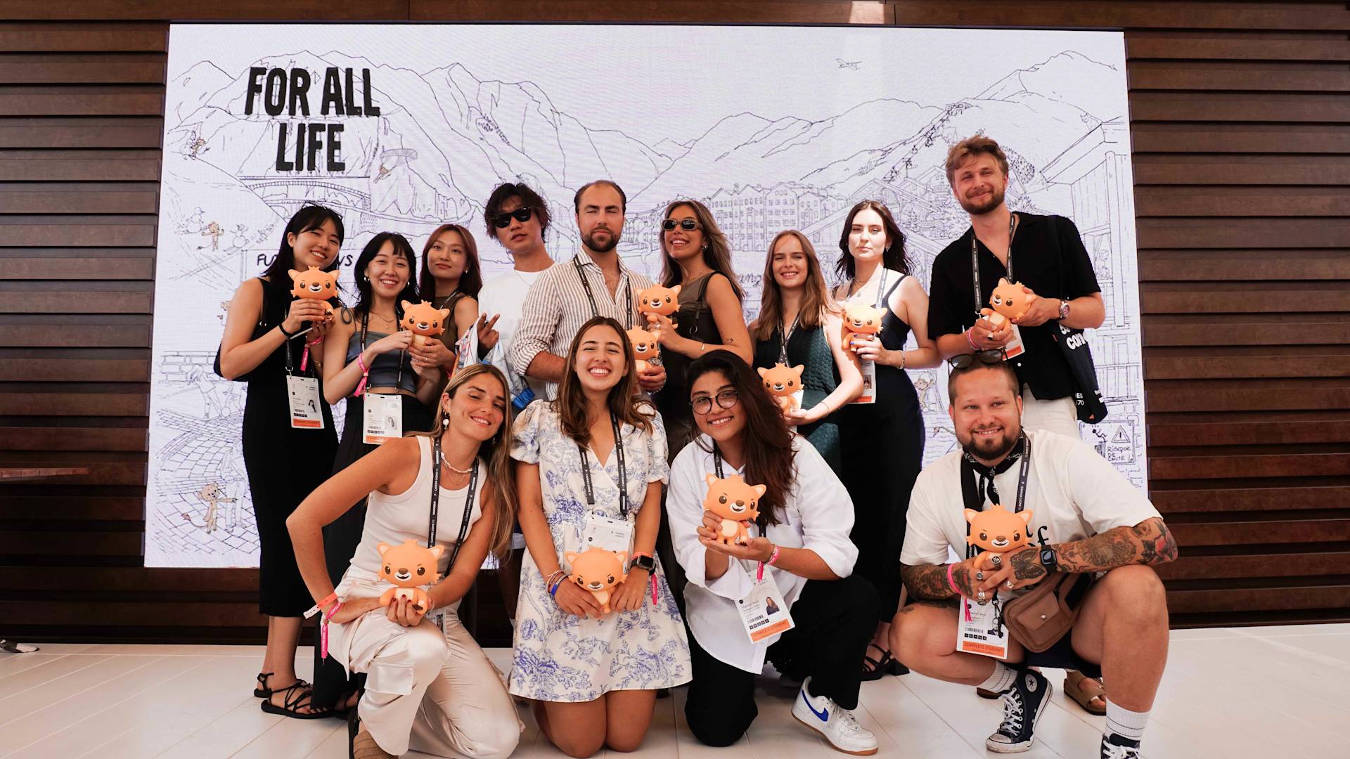 A group of people pose and smile at the camera, each holding a small orange cat figurine. They stand in front of a large backdrop with a mountain illustration and the words “FOR ALL LIFE.”.