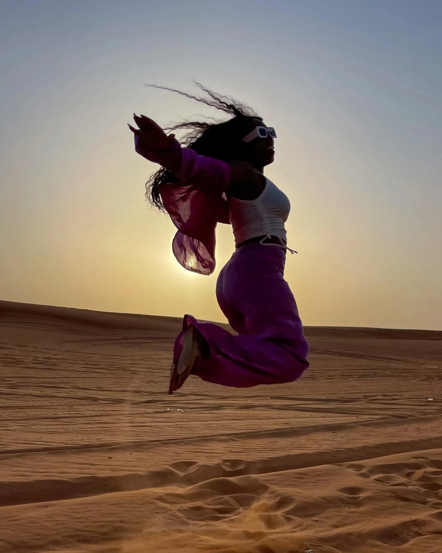 A woman wearing sunglasses, a white top, and purple outfit jumps joyfully in a desert at sunset, with her hair and clothes flowing in the air and sand beneath her.