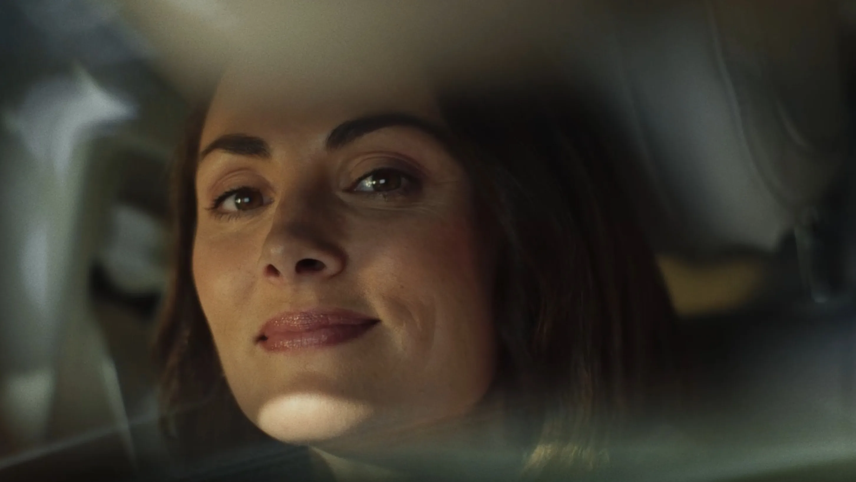 A woman with dark hair and brown eyes sits in a car, looking out the window with a slight, confident smile. The image is softly focused with warm lighting on her face.