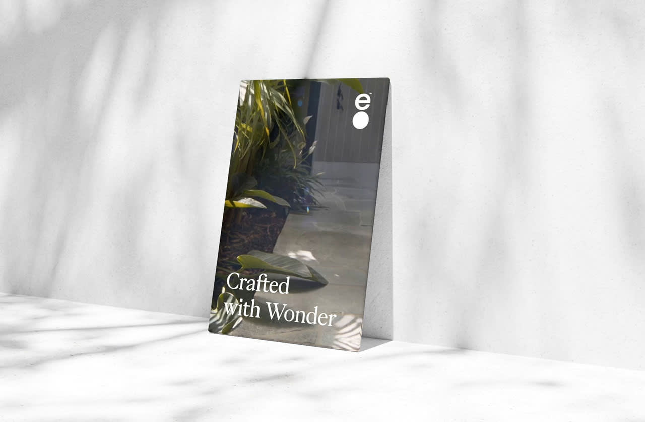 A poster leaning against a white wall with the text Crafted With Wonder. The poster shows green plants and a walkway, and soft shadows fall across the scene.