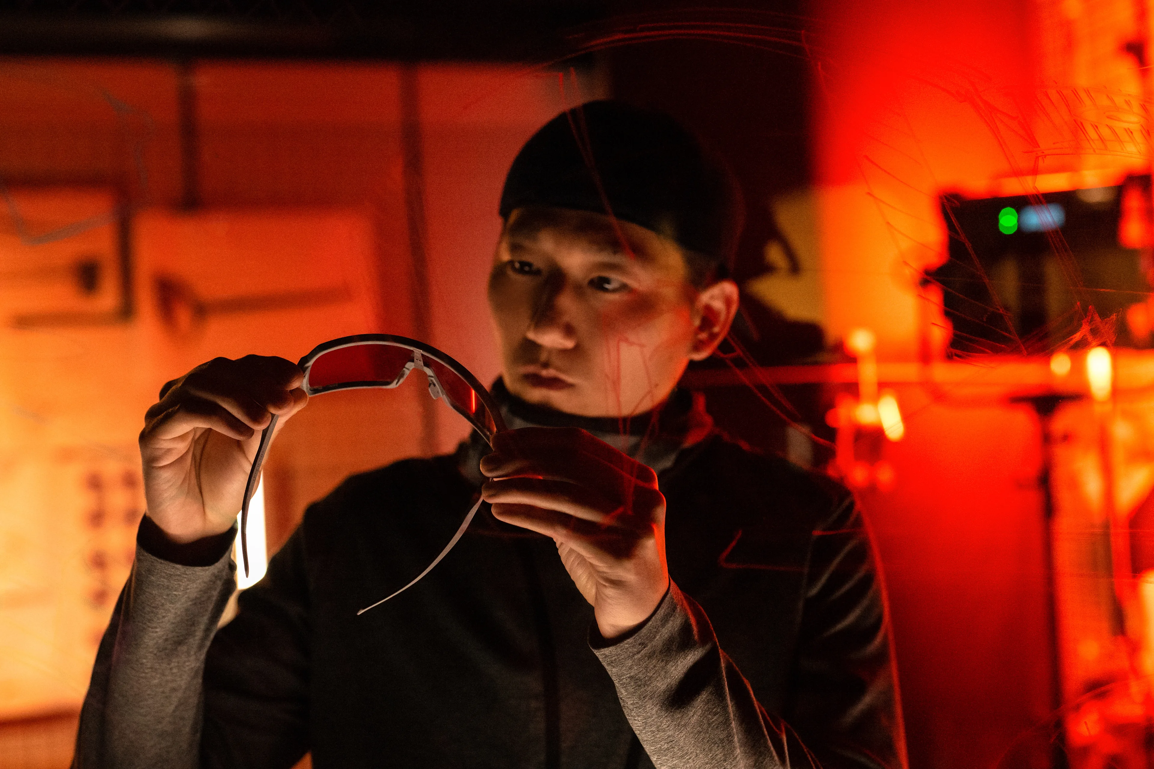 A person in a dark room with red lighting holds and examines a pair of tinted glasses, looking at them closely. Technical equipment and papers are visible in the background.