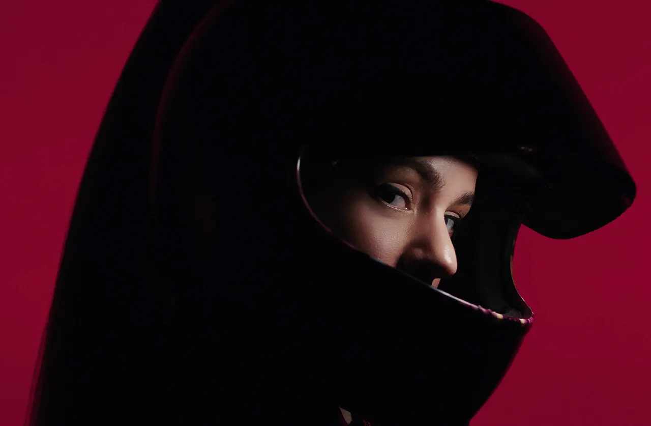 A person wearing a black motorcycle helmet looks to the side against a deep red background, with only their eyes and part of their face visible through the helmets opening.