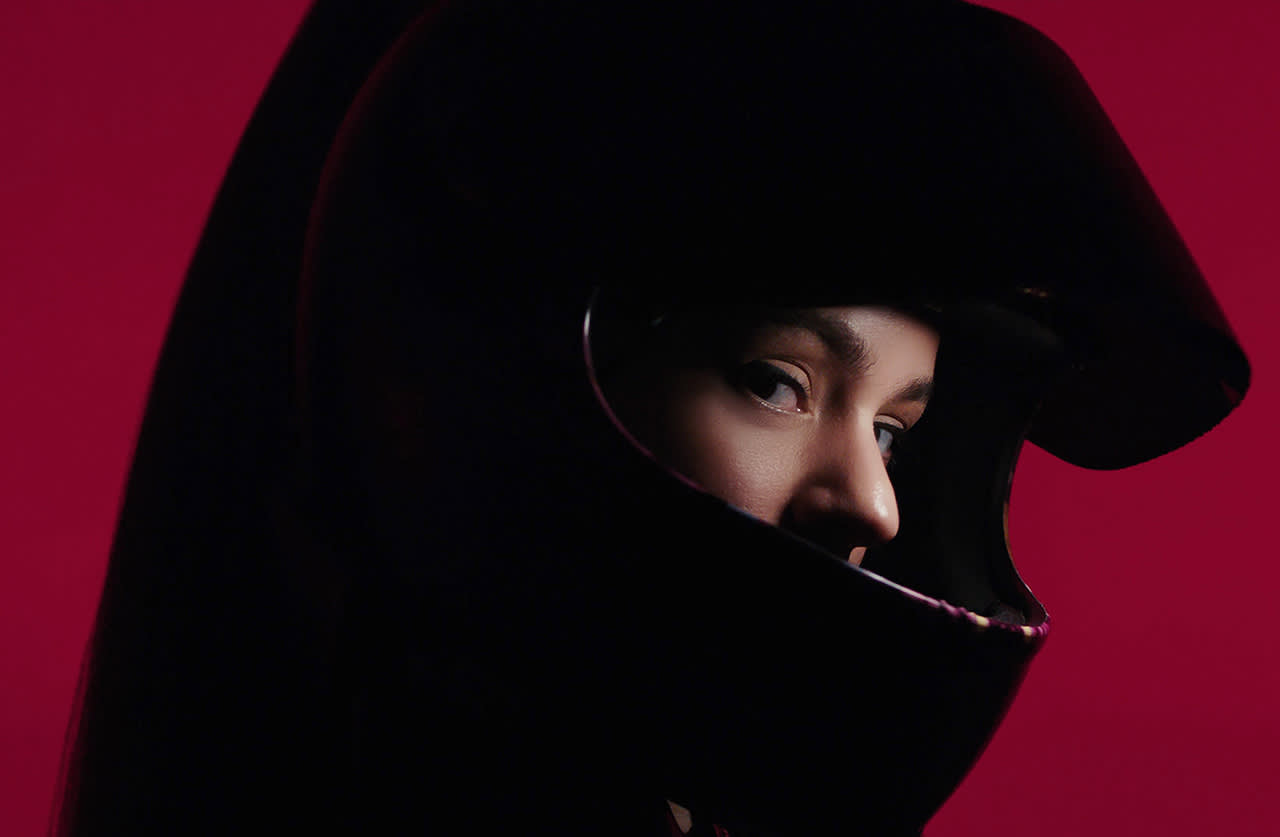 A person wearing a black motorcycle helmet looks to the side against a deep red background, with only their eyes and part of their face visible through the helmets opening.
