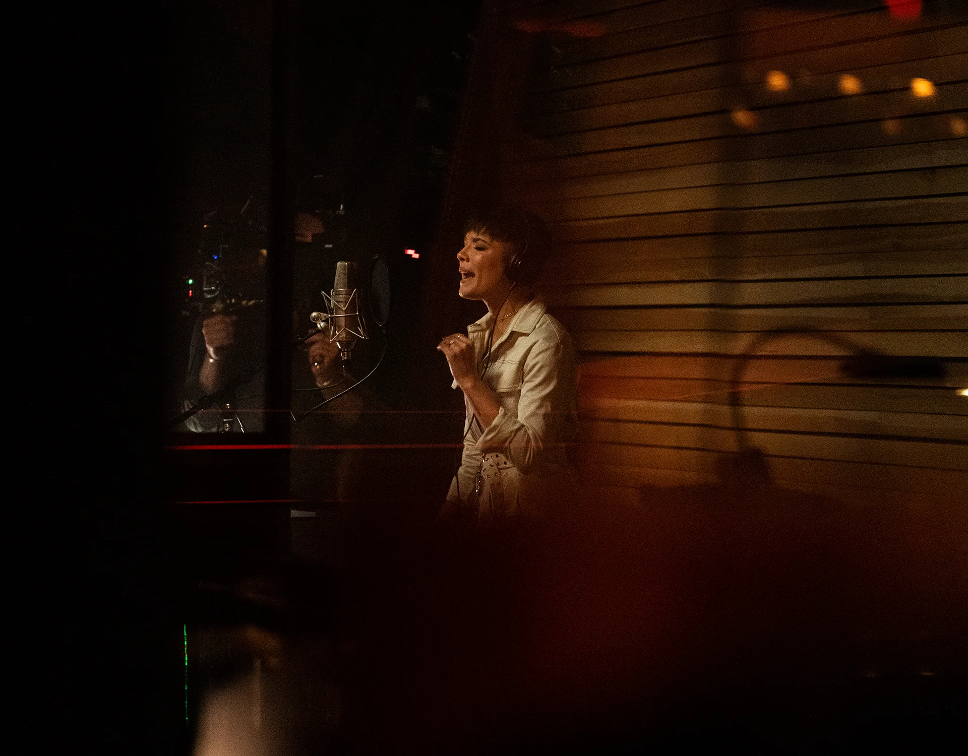 A woman in a white shirt sings passionately into a studio microphone in a dimly lit recording booth with wooden panel walls.