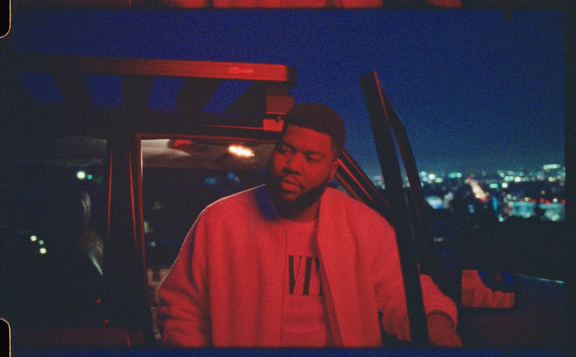 A man stands by an open car door at night, illuminated by red and blue lights, with a cityscape glowing in the background. He wears a light jacket and looks thoughtfully to the side.