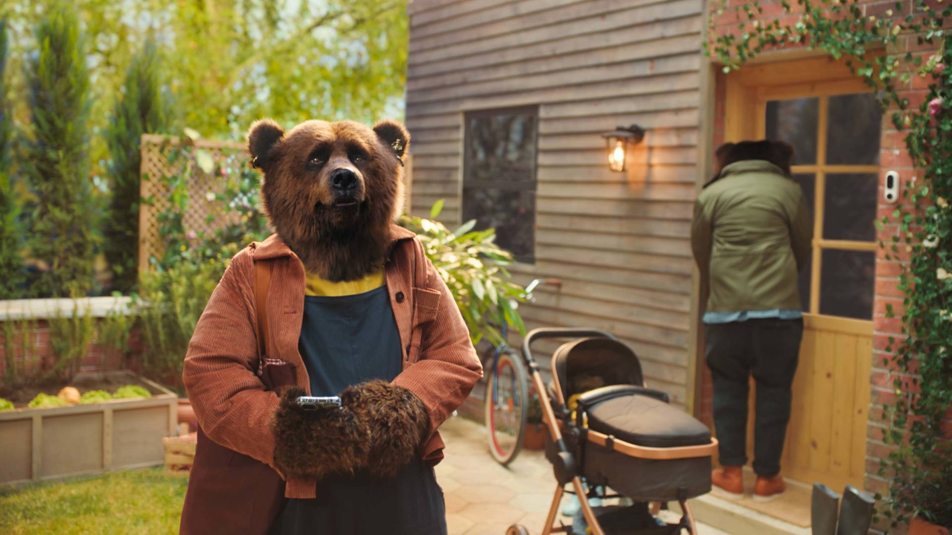 A bear dressed in human clothes stands outside a house holding a smartphone, with a stroller and bicycle nearby. Another bear, also dressed in clothes, is entering the house through the door.