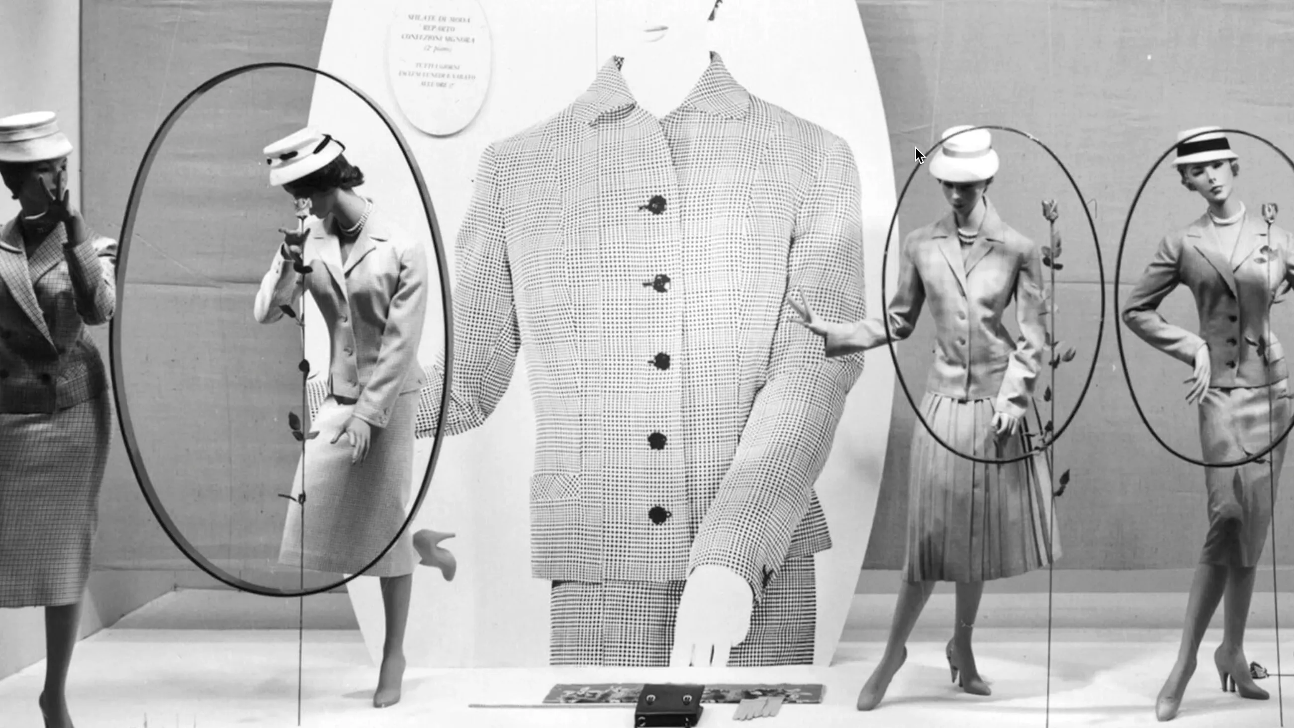 Three mannequins in vintage suits and hats pose with roses in a store display, with a large-scale jacket and hand illustration as the backdrop. Ovals frame the mannequins, creating an elegant composition.