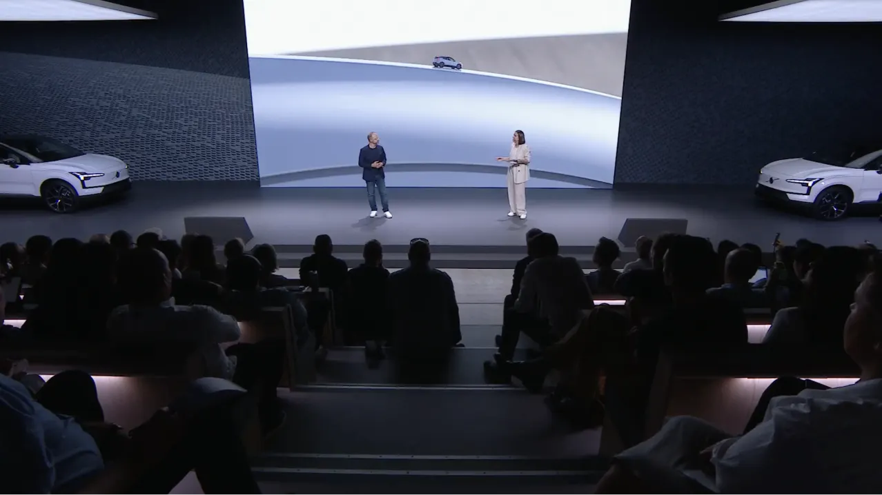 Two people stand on stage presenting to an audience, with large screens behind them displaying a futuristic car and abstract shapes; two white cars are partially visible on either side of the stage.