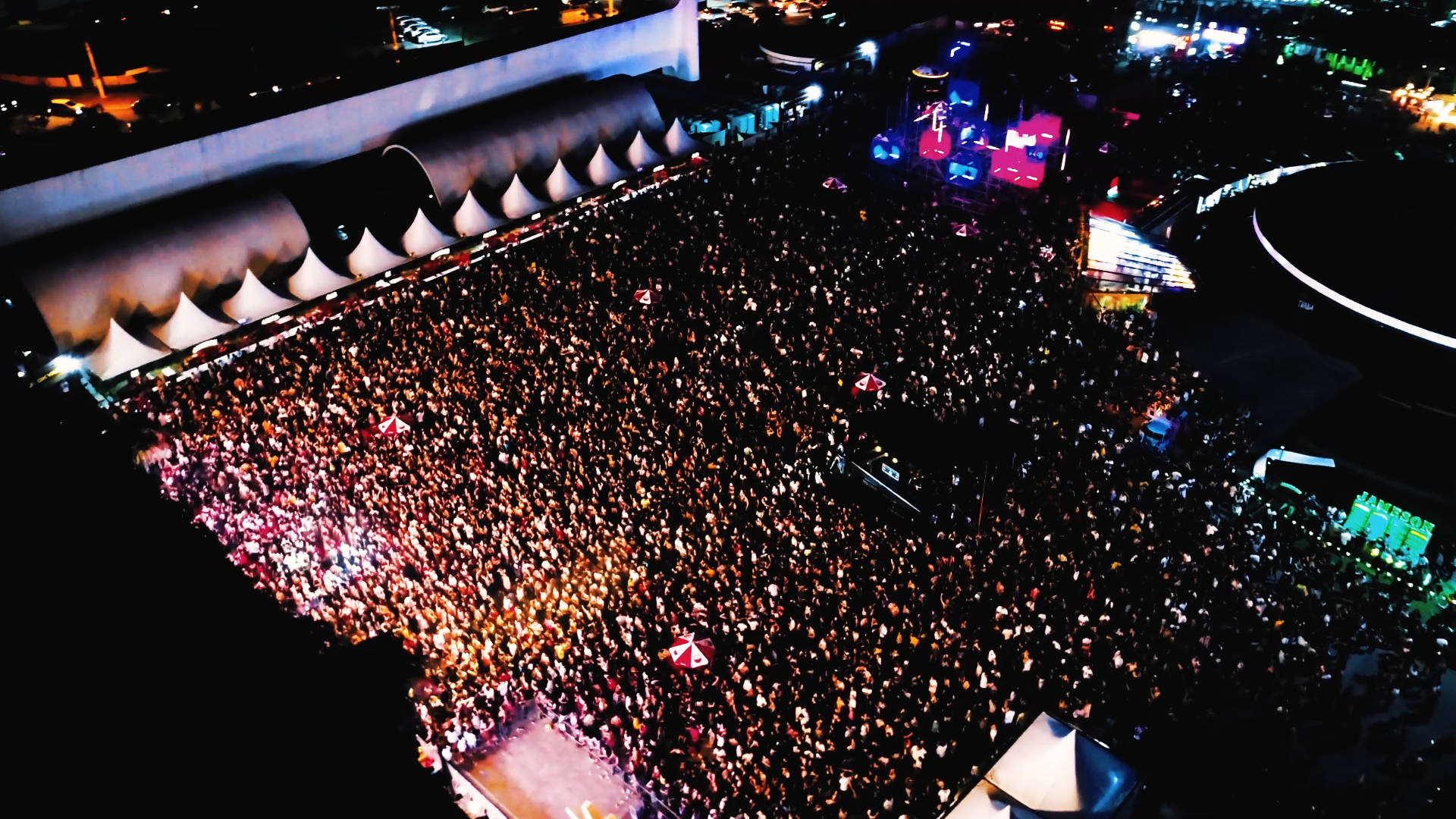 A large crowd gathers outdoors at night for a concert or festival, filling the area in front of a brightly lit stage with colorful lights and screens. Tents line one side of the venue.