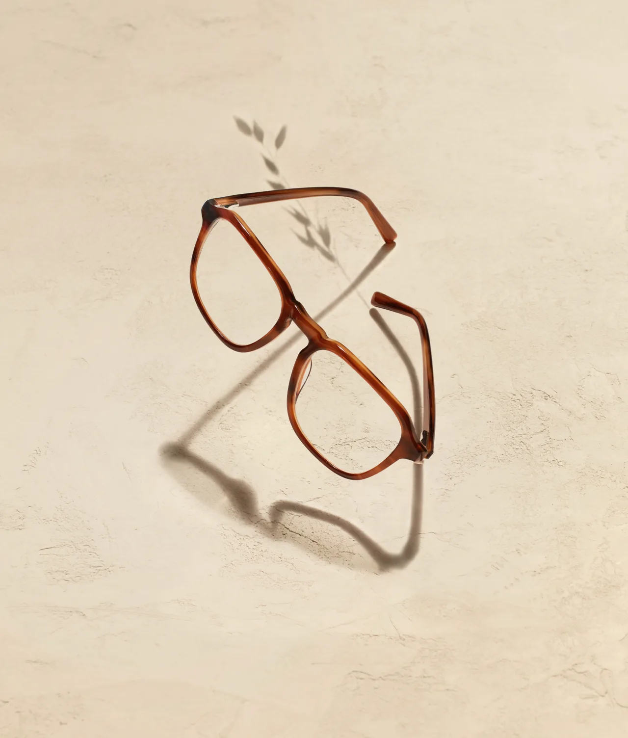 A pair of brown eyeglasses with large square frames rests on a textured beige surface, casting a distinct shadow and a small branch-shaped shadow nearby.