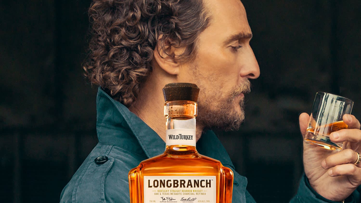 A man with curly hair and a beard in profile holds a glass of whiskey. In the foreground, a bottle of Wild Turkey Longbranch bourbon whiskey is prominently displayed.