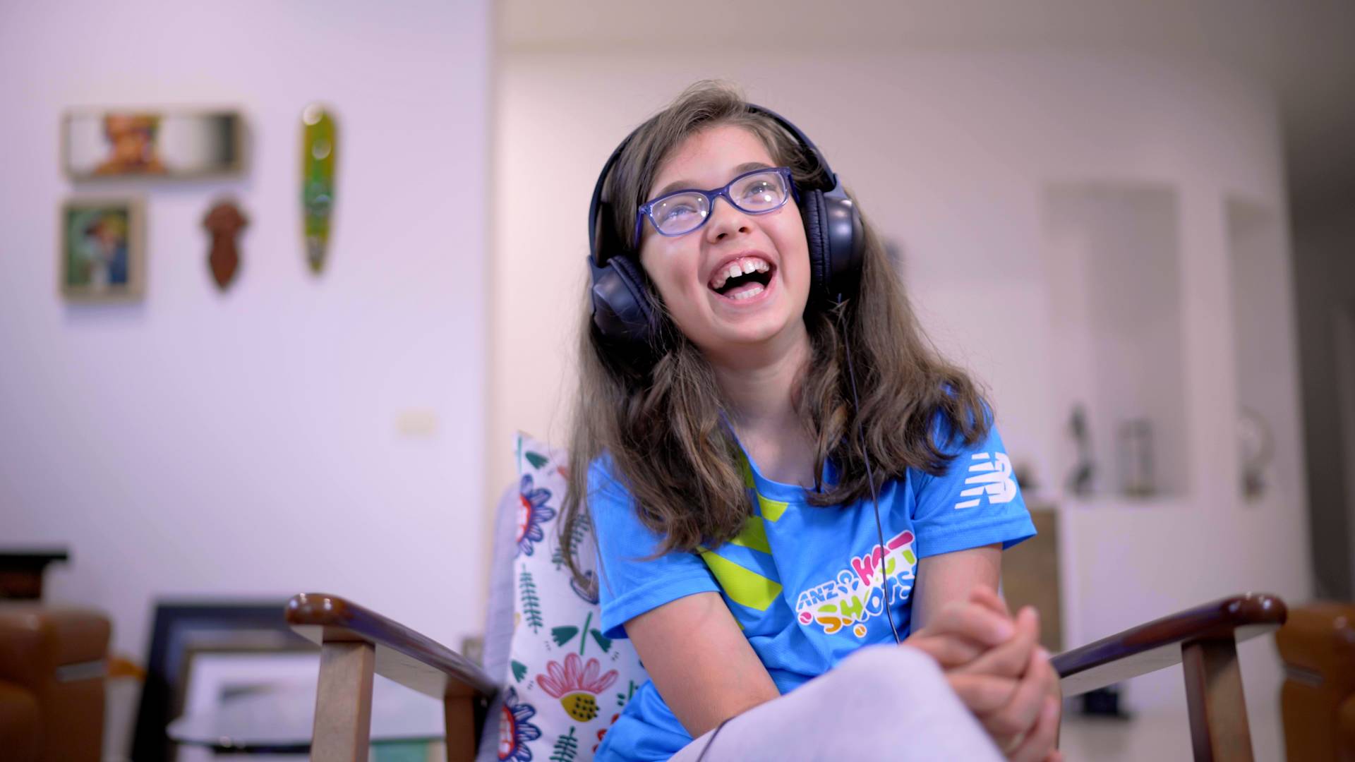 A young girl with long brown hair and glasses sits in a chair, smiling and laughing while wearing large headphones. She is dressed in a blue T-shirt and light pants, appearing happy and relaxed indoors.