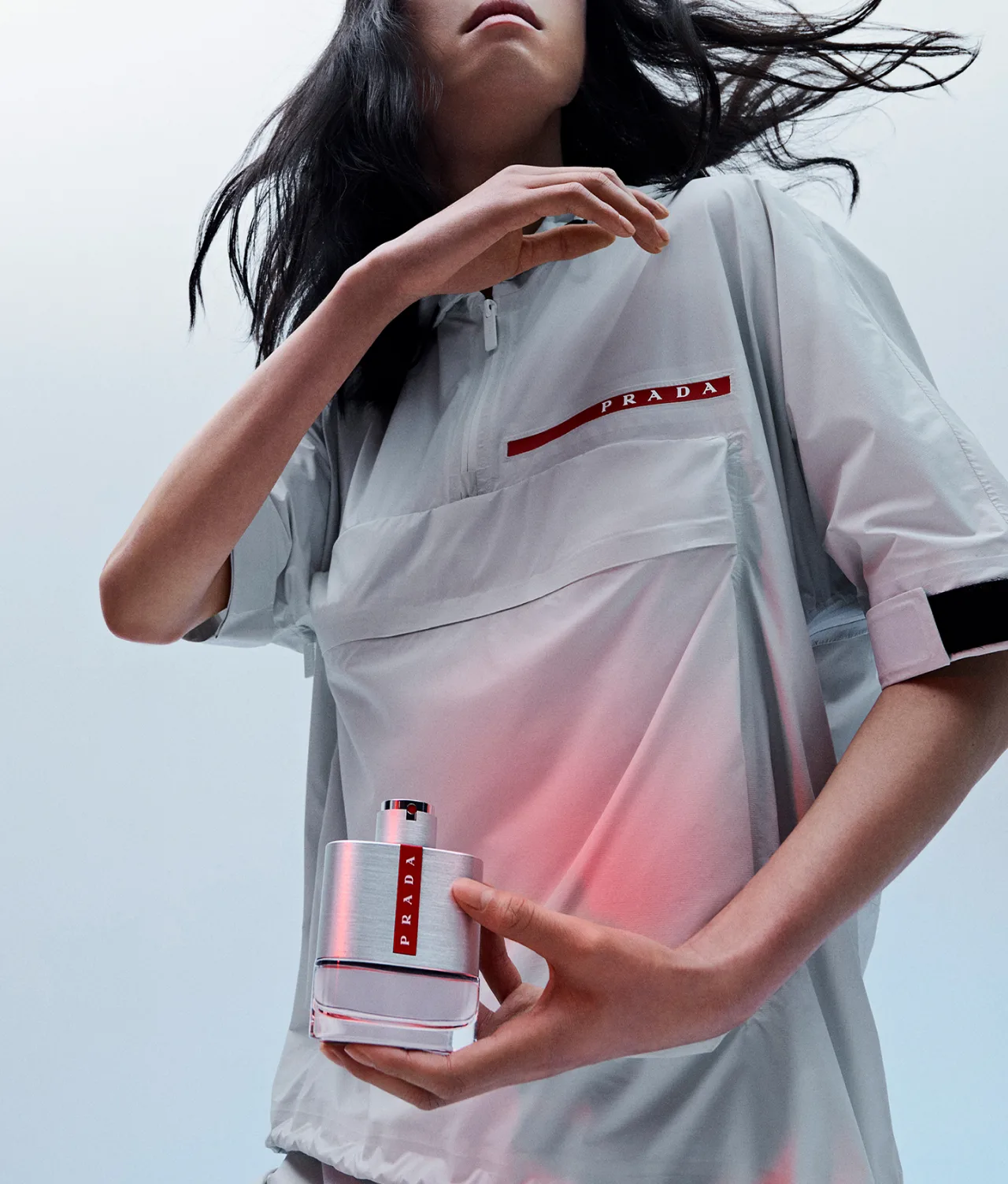 A person in a light gray Prada shirt holds a silver and red Prada perfume bottle. Their hair is loose, and the image has a cool, modern aesthetic with soft lighting.