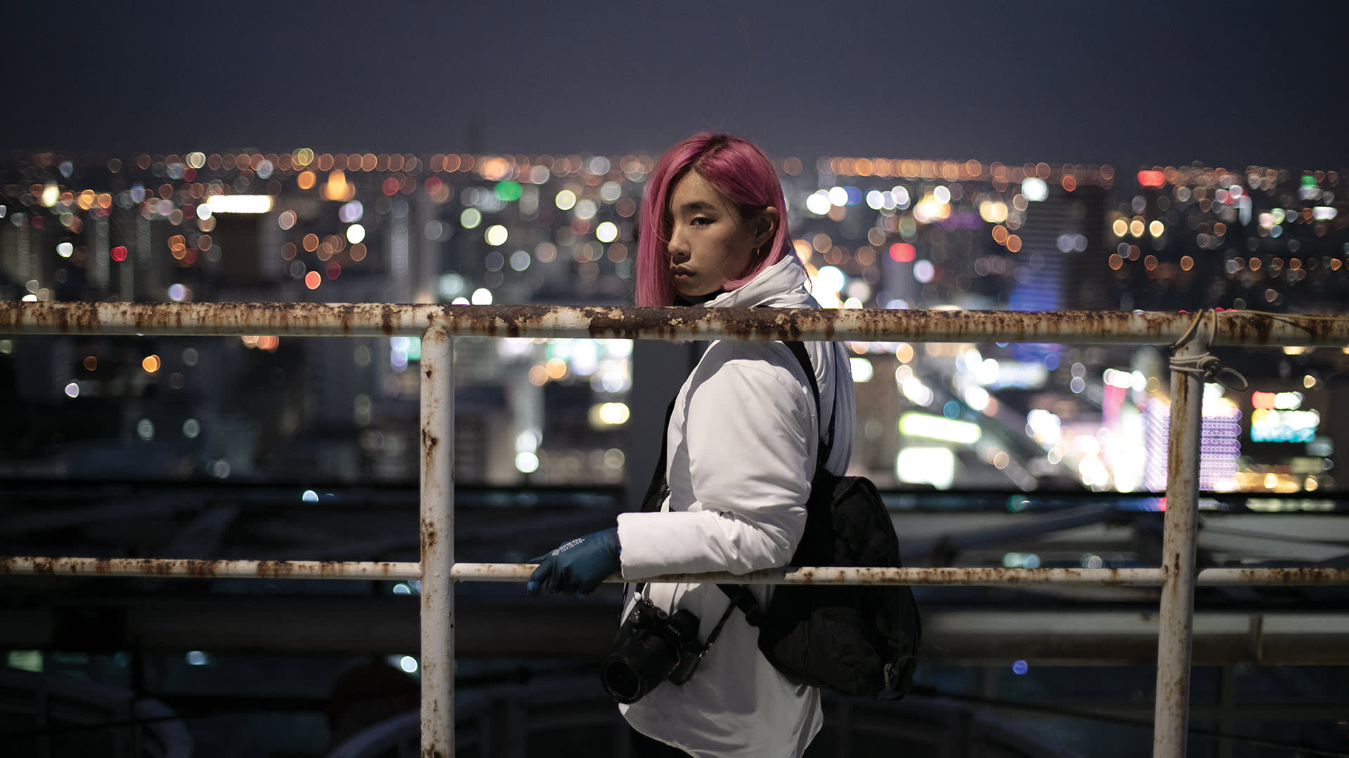 A woman with pink hair standing on a railing with a city in the background.