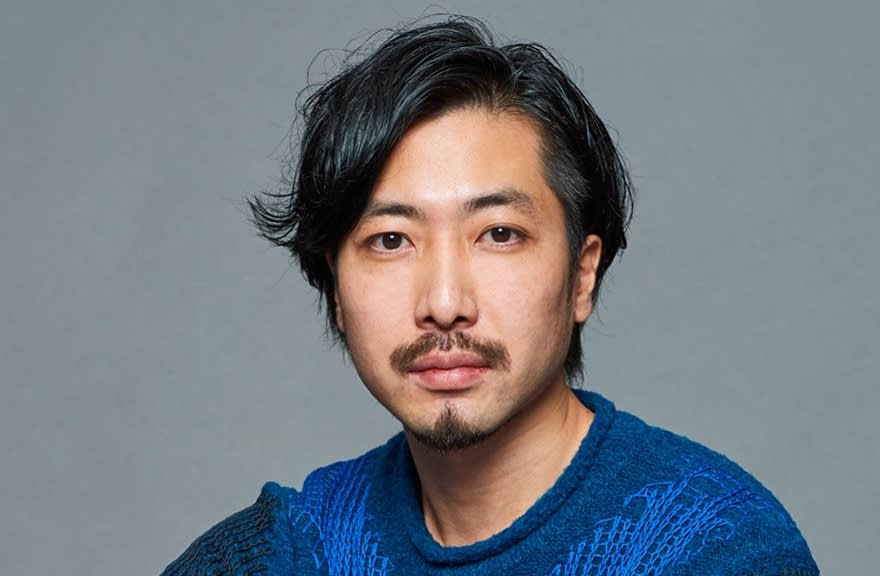 A man with medium-length black hair, a mustache, and a short beard is wearing a textured blue sweater and looking directly at the camera against a plain gray background.