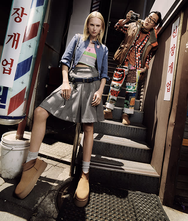 A woman in a grey skirt and oversized tan shoes stands on stairs, while a man in colorful clothing behind her holds a camera. They are outside, between buildings with Korean signs.