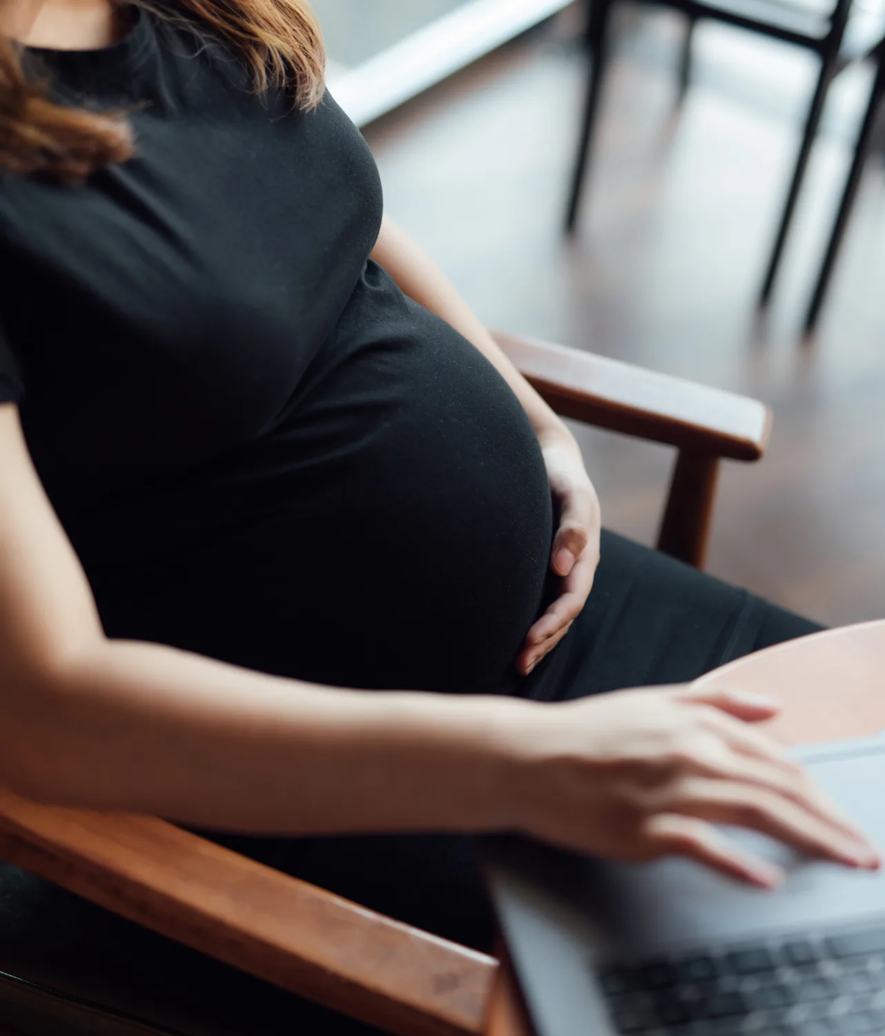 A pregnant person in a black dress sits in a chair, resting one hand on their belly and the other hand on a laptop. The photo is taken indoors, capturing a calm and focused moment.