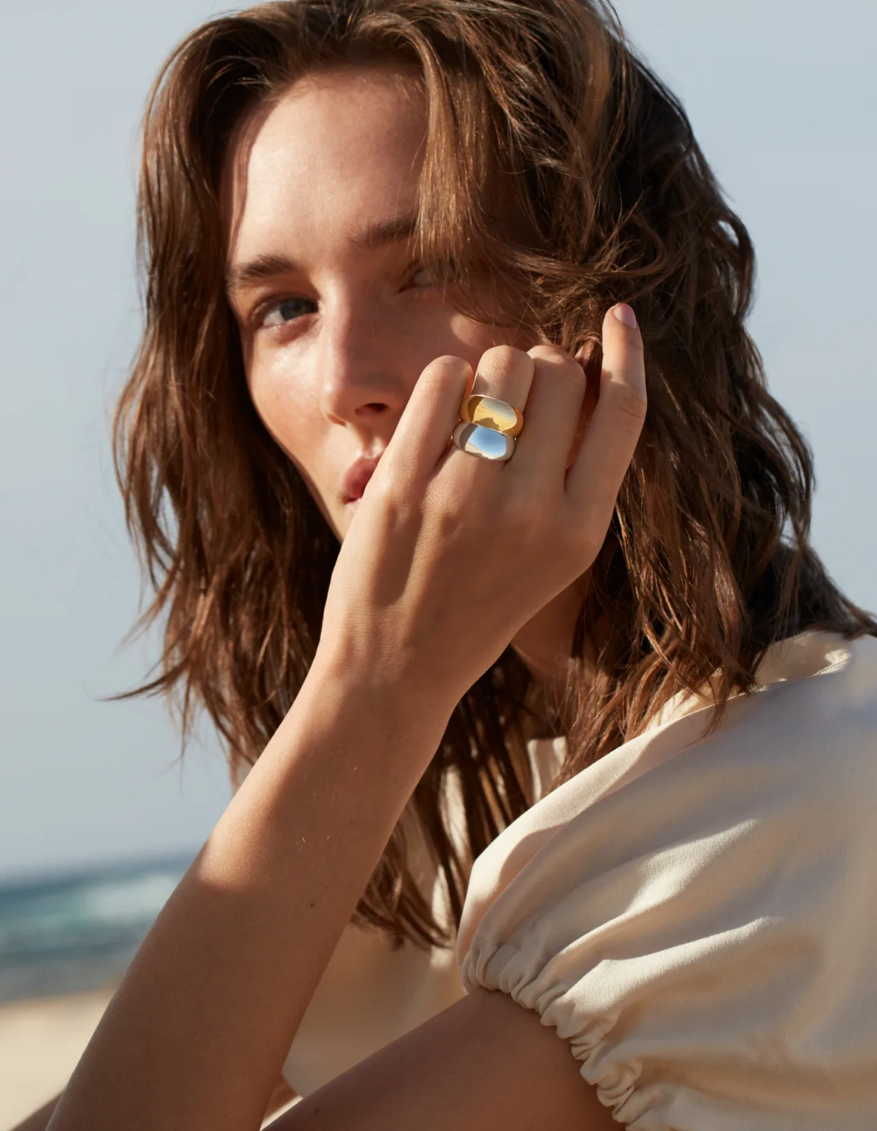 A woman with wavy brown hair wears a beige blouse and holds her hand near her face, showcasing a large gold ring with a white stone. She is outdoors, with the beach and sea visible in the blurred background.