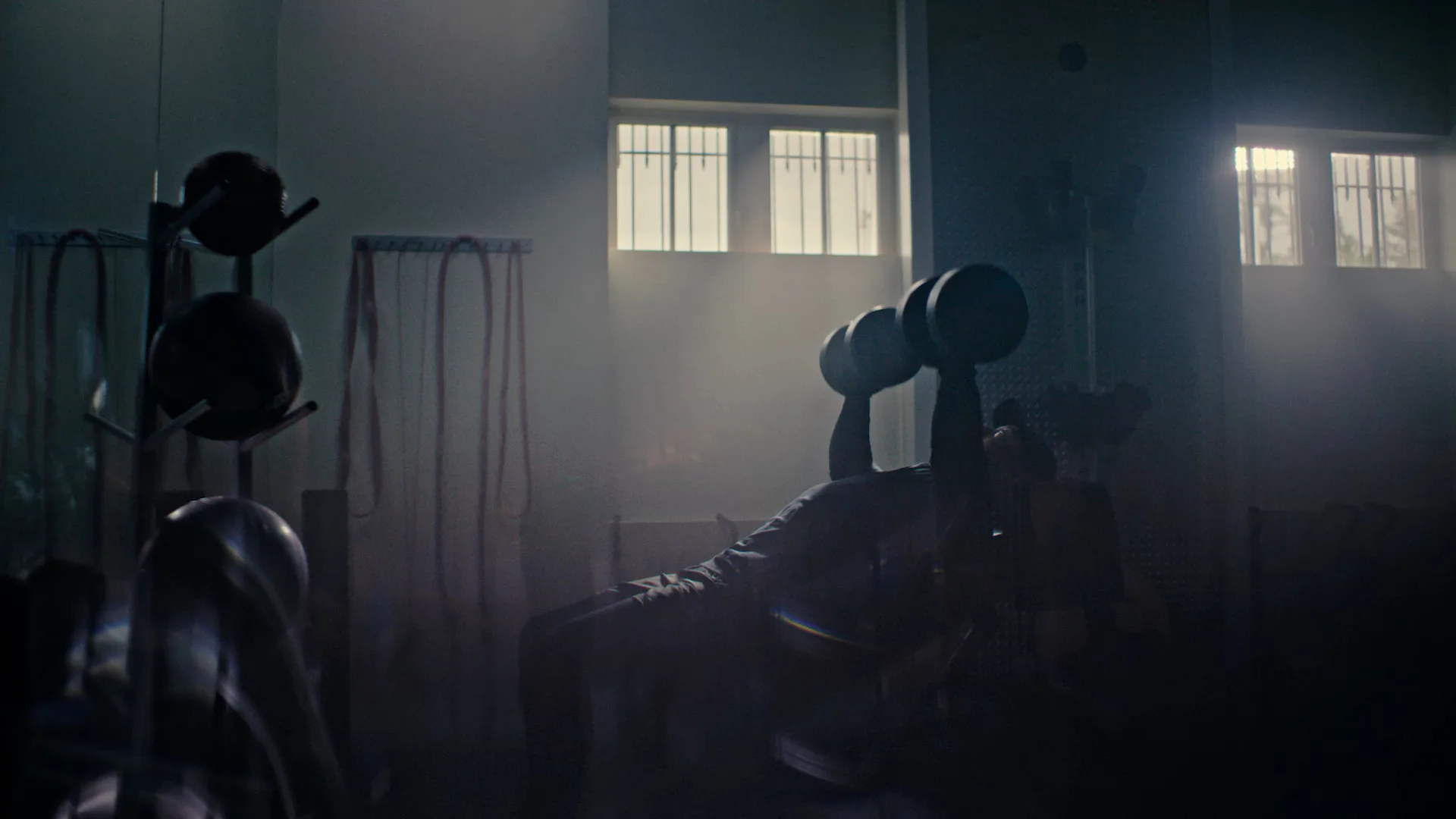A person lifts dumbbells while lying on a bench in a dim, shadowy gym. Light from a window highlights the misty or dusty air, and gym equipment is visible in the background.