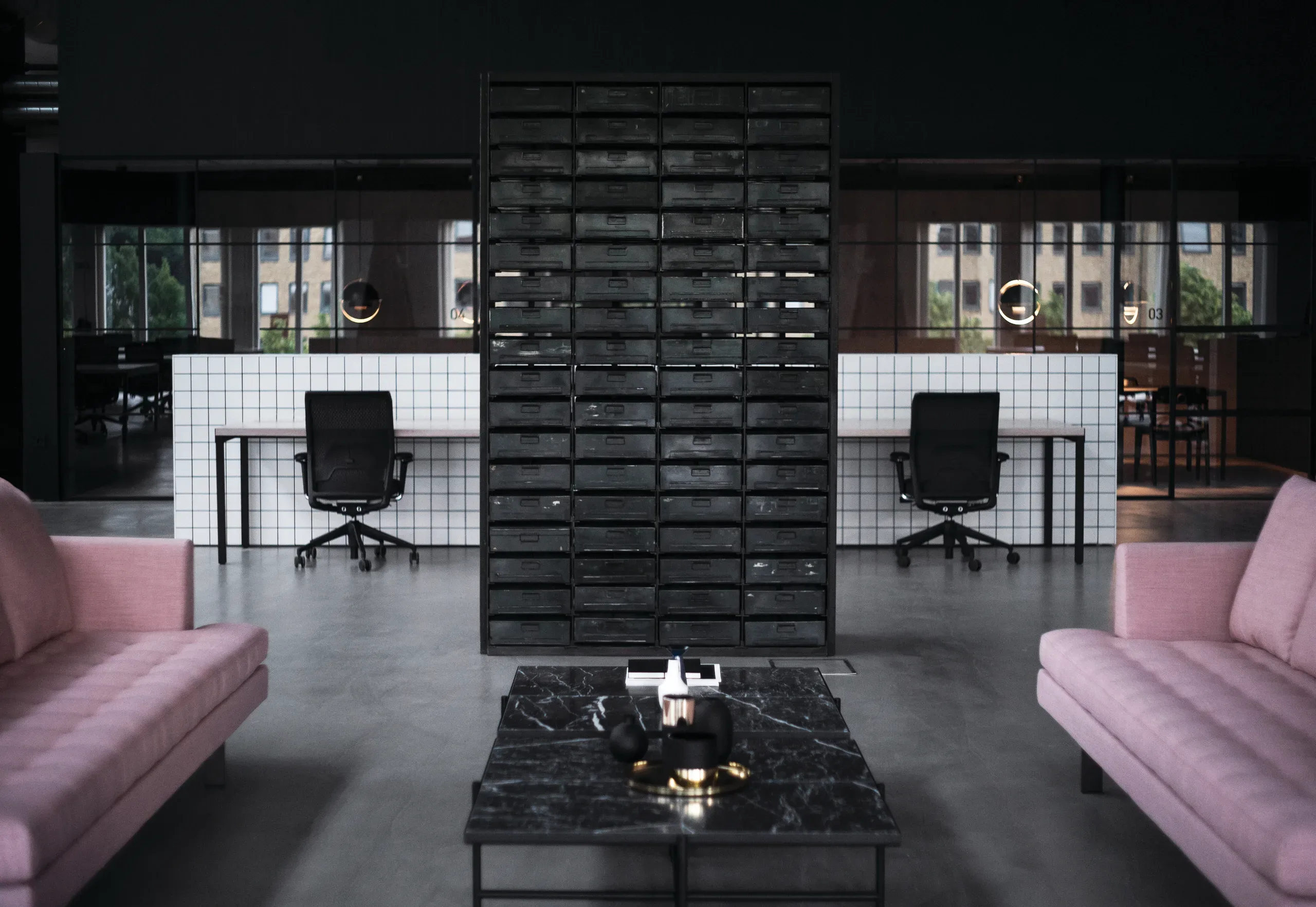 Modern office space with two pink sofas, a black marble coffee table, a large black shelving unit, and two white desks with office chairs in the background. Large windows let in natural light.