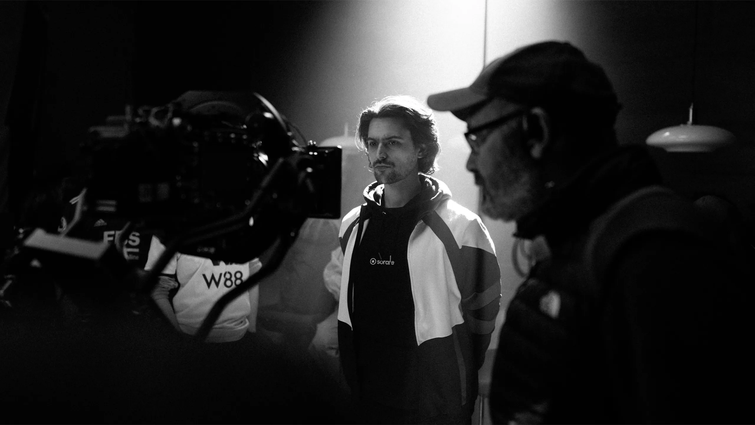 A black and white photo showing a man in a hoodie standing under dramatic lighting, surrounded by film equipment and crew, with a camera in the foreground and another person in a cap nearby.