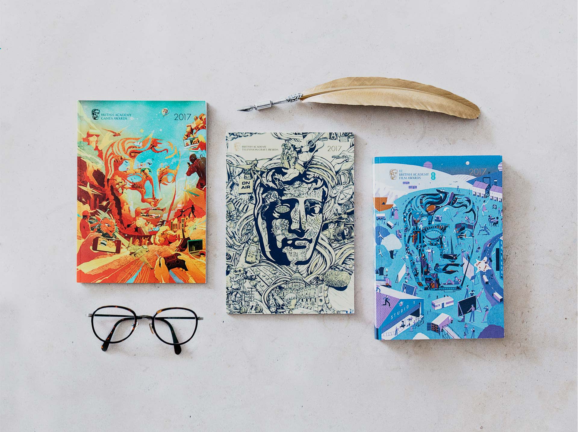 Three notebooks with artistic covers featuring a stylized face, a quill pen, and a pair of black eyeglasses are arranged on a light surface. The covers are colorful, with one in orange, one in blue, and one in black and white.