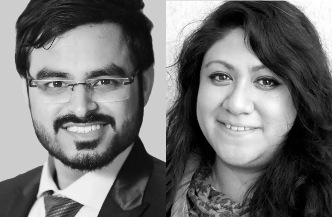 Two people side by side: on the left, a man with glasses, a beard, and a suit, smiling; on the right, a woman with long wavy hair, also smiling. The image is in black and white.