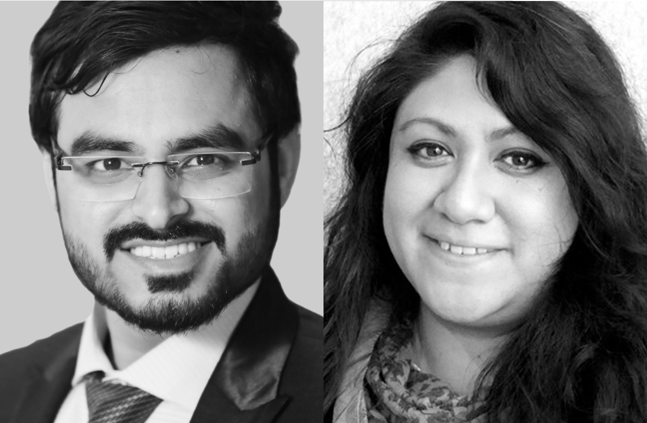 Two people side by side: on the left, a man with glasses, a beard, and a suit, smiling; on the right, a woman with long wavy hair, also smiling. The image is in black and white.