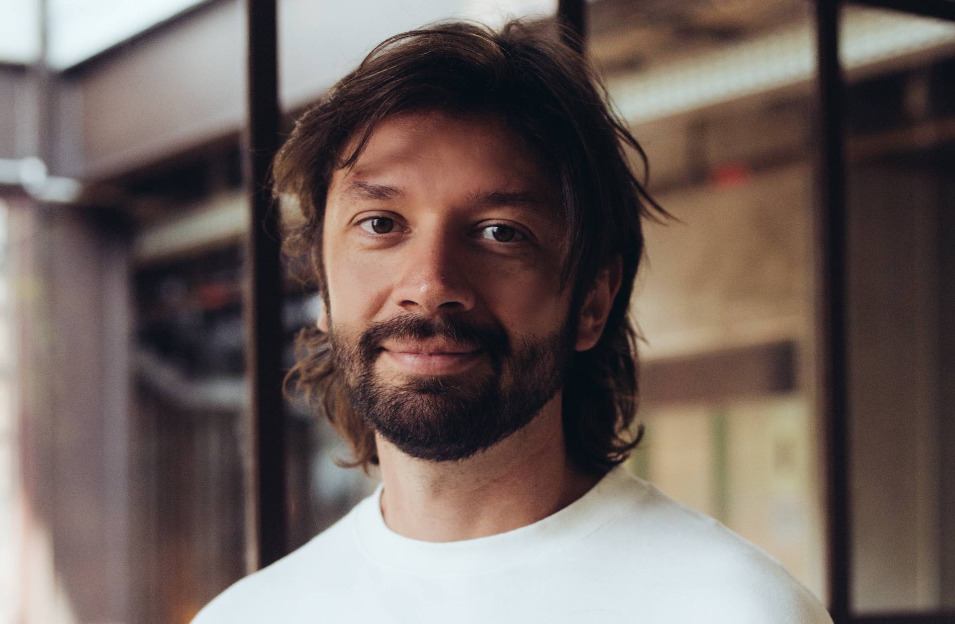 A man with medium-length brown hair and a beard, wearing a white shirt, standing indoors near large windows with a soft smile and blurred background.