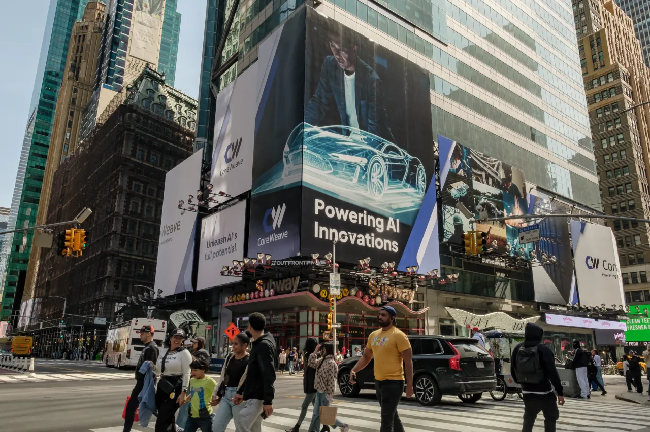 People cross a busy intersection in Times Square, New York City, with large digital billboards displaying advertisements about AI innovations and electric vehicles on nearby buildings. Cars and tall skyscrapers surround the scene.