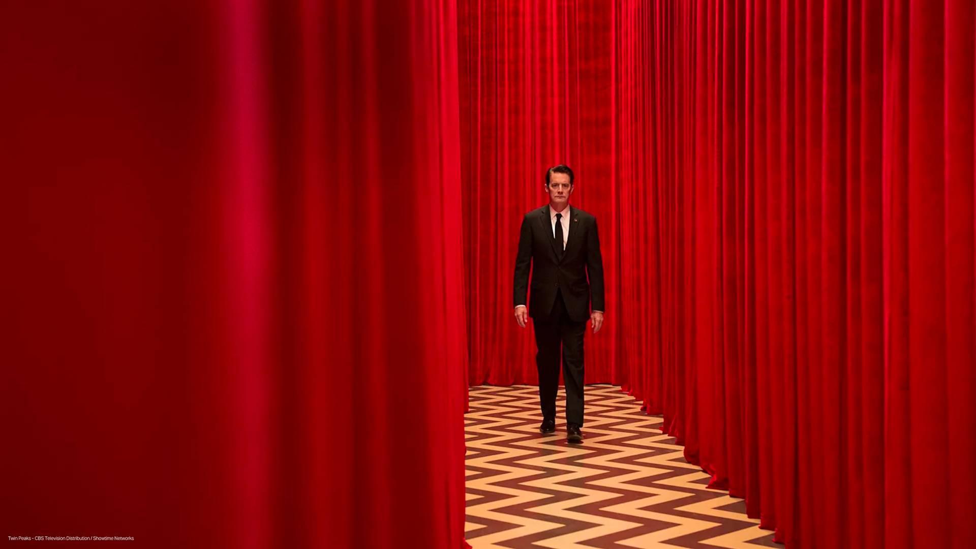 A man in a black suit walks through a corridor with red curtains and a black-and-white zigzag patterned floor. The atmosphere is surreal and dramatic.