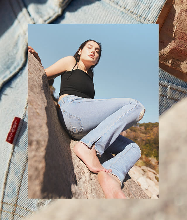 A person in a black sleeveless top and light blue jeans sits barefoot on a large rock outdoors, with a clear blue sky and greenery in the background. The image is framed by a denim fabric border.