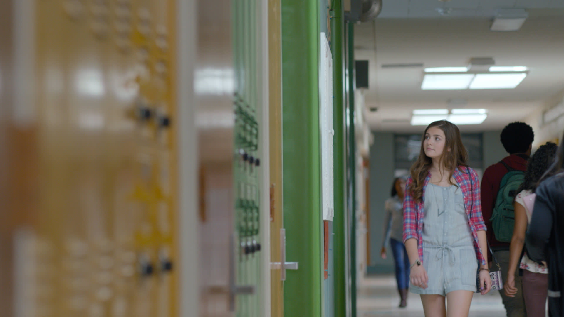 A teenage girl wearing a plaid shirt over a light dress walks down a school hallway lined with lockers, looking to her left. Other students are visible in the background.