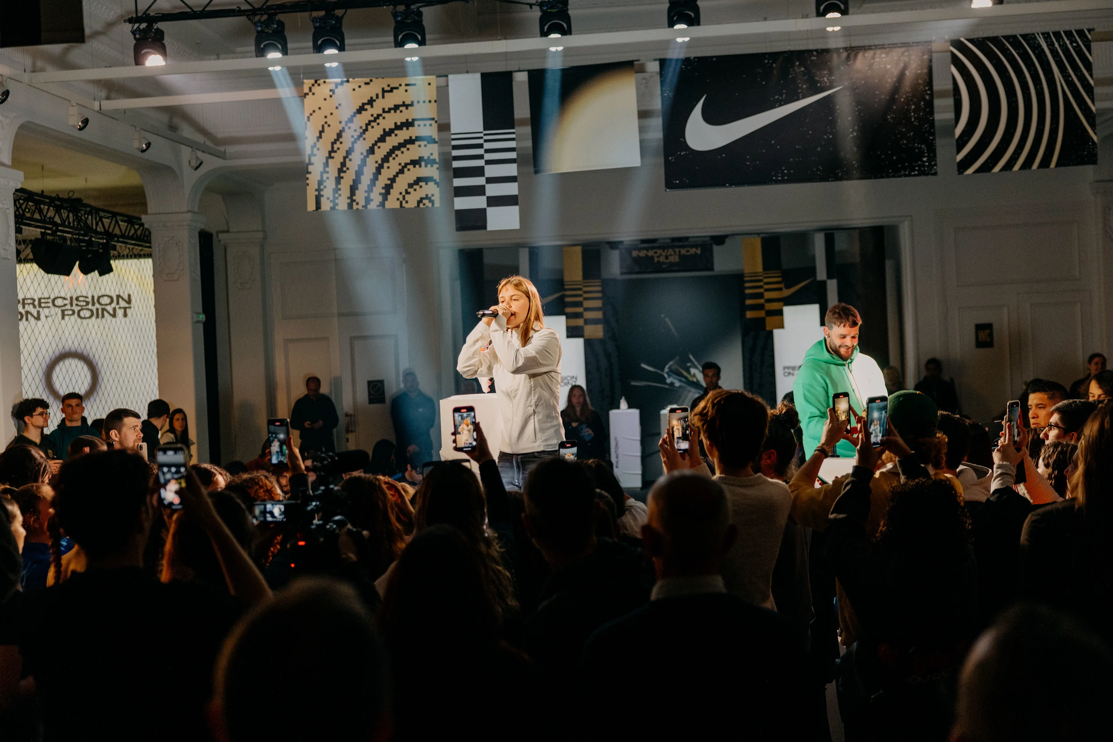 A person in a white hoodie sings on stage at an indoor event, surrounded by a crowd taking photos. Branded banners, including Nike, hang above, and stage lights shine down on the scene.