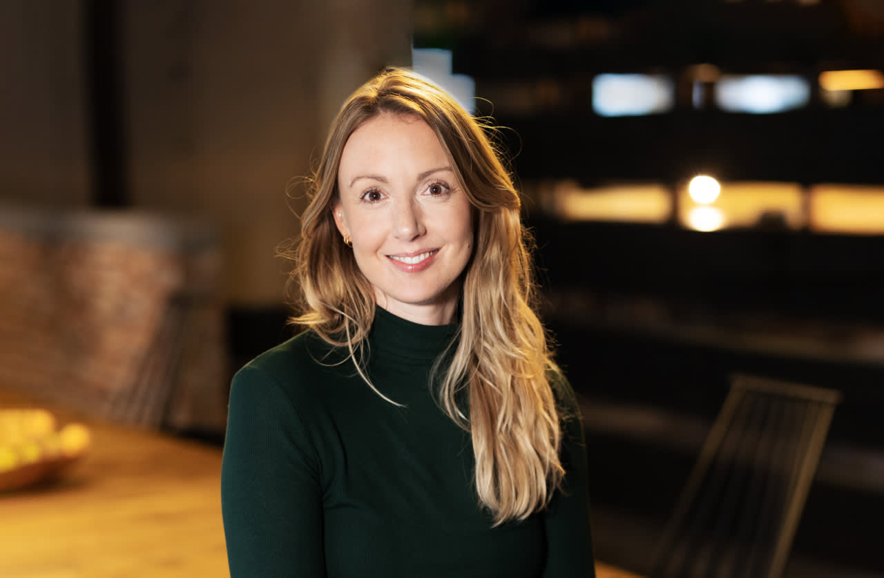 A woman with long blonde hair wearing a dark green turtleneck sweater is smiling at the camera. She is in a warmly lit indoor setting with blurred background elements.
