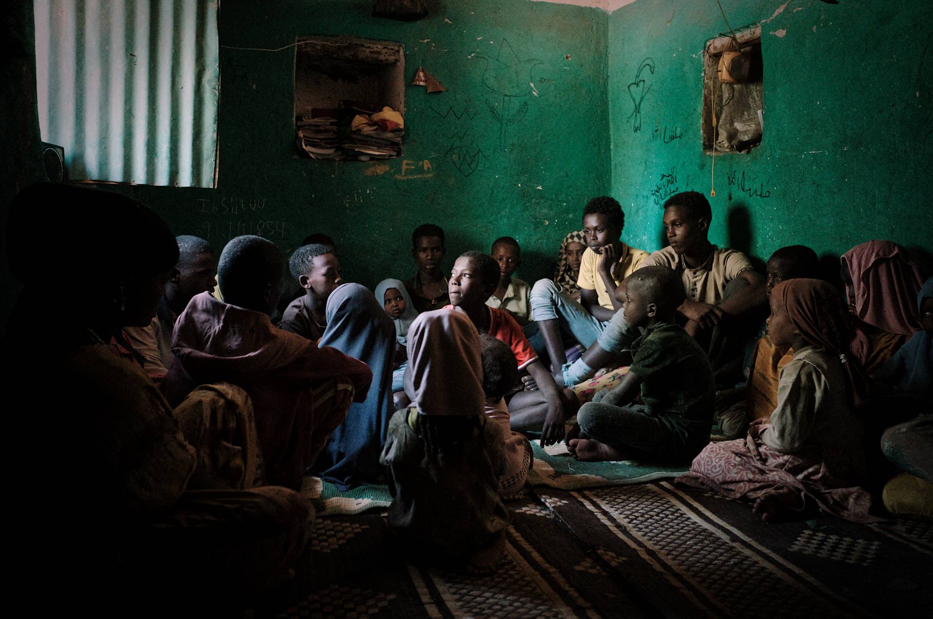 A group of children and young adults sit closely together on the floor of a dimly lit room with green walls, paying attention to someone out of view. Light enters through a window, highlighting their attentive faces.