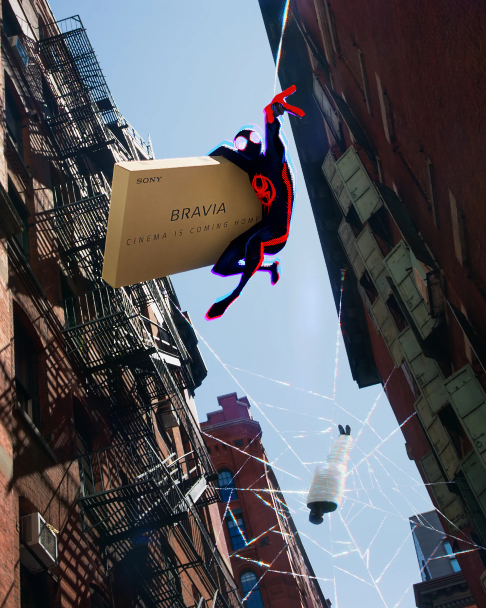 A Spider-Man figure swings between brick buildings, holding a large Sony Bravia TV box attached to a web. In the background, a spool of thread hangs in a web against a blue sky.