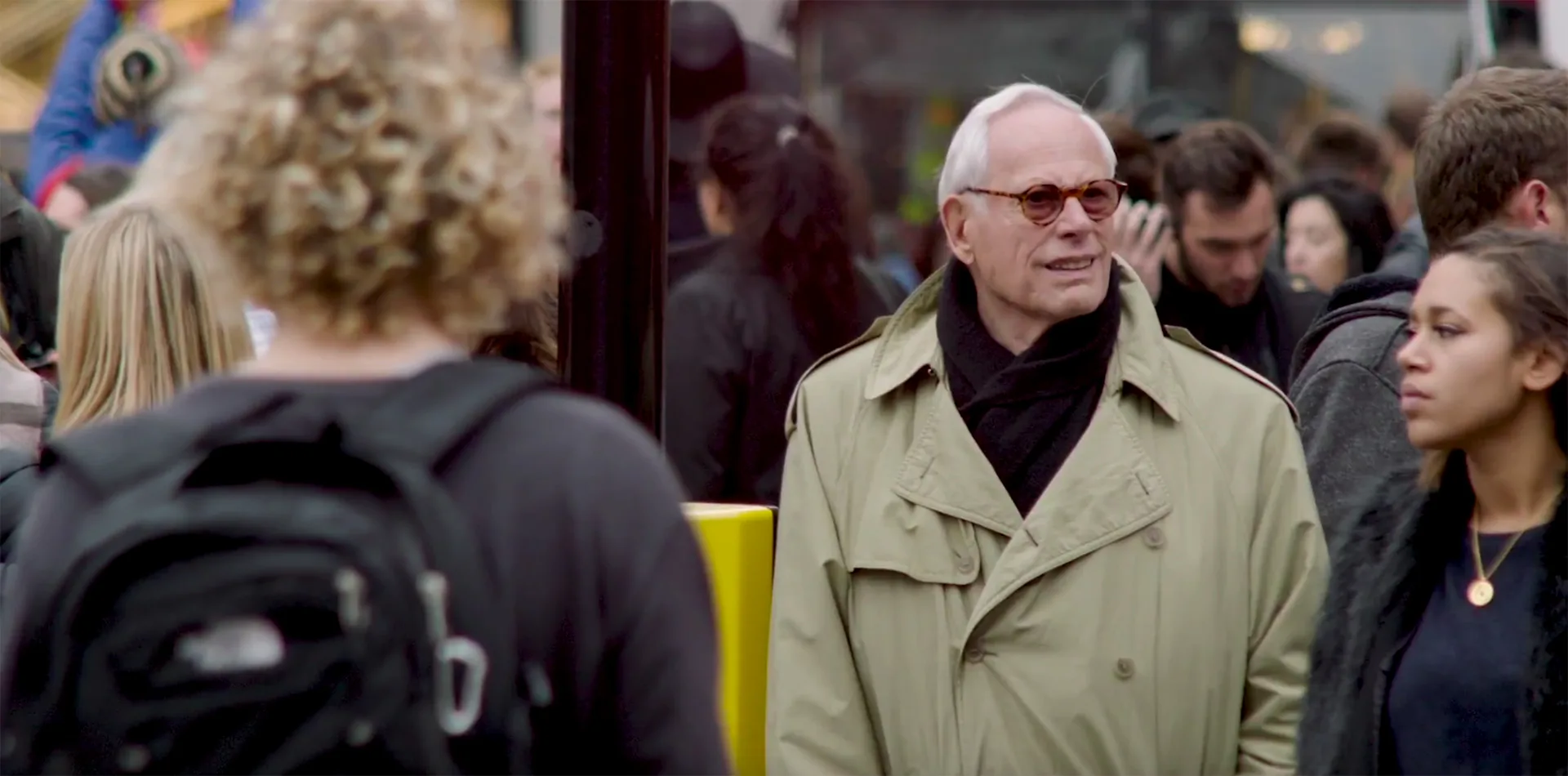 An older man in a beige trench coat and sunglasses stands among a crowd of people on a city street, looking to the side. Several people are visible around him, including a young woman and a person with a backpack.