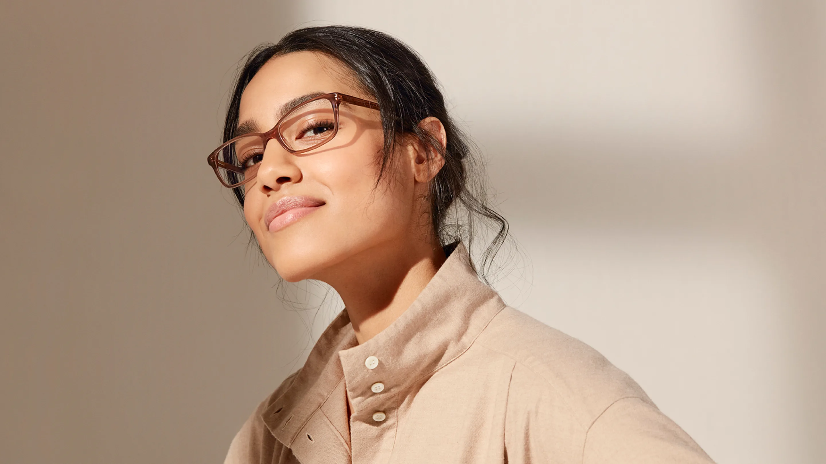 A woman wearing brown glasses and a beige collared shirt looks confidently at the camera, with her dark hair pulled back and a subtle smile on her face, against a softly lit neutral background.
