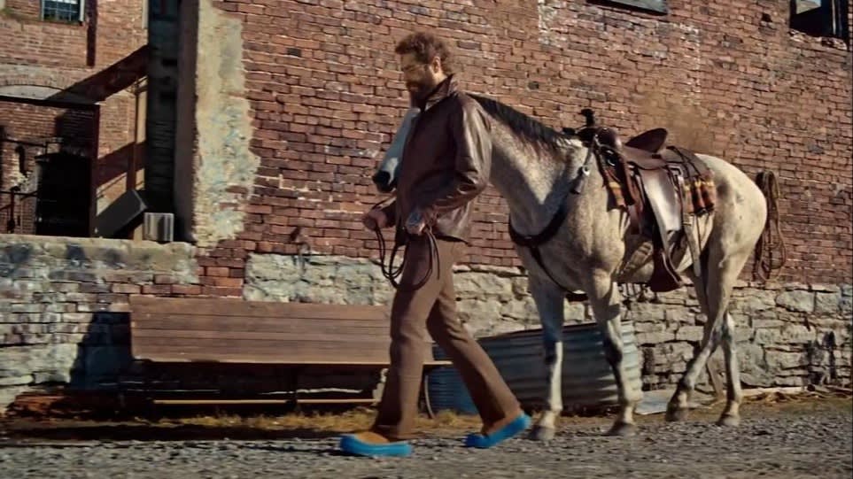 A man wearing a brown jacket and blue shoes walks beside a saddled gray horse along a brick building with a wooden bench outside.
