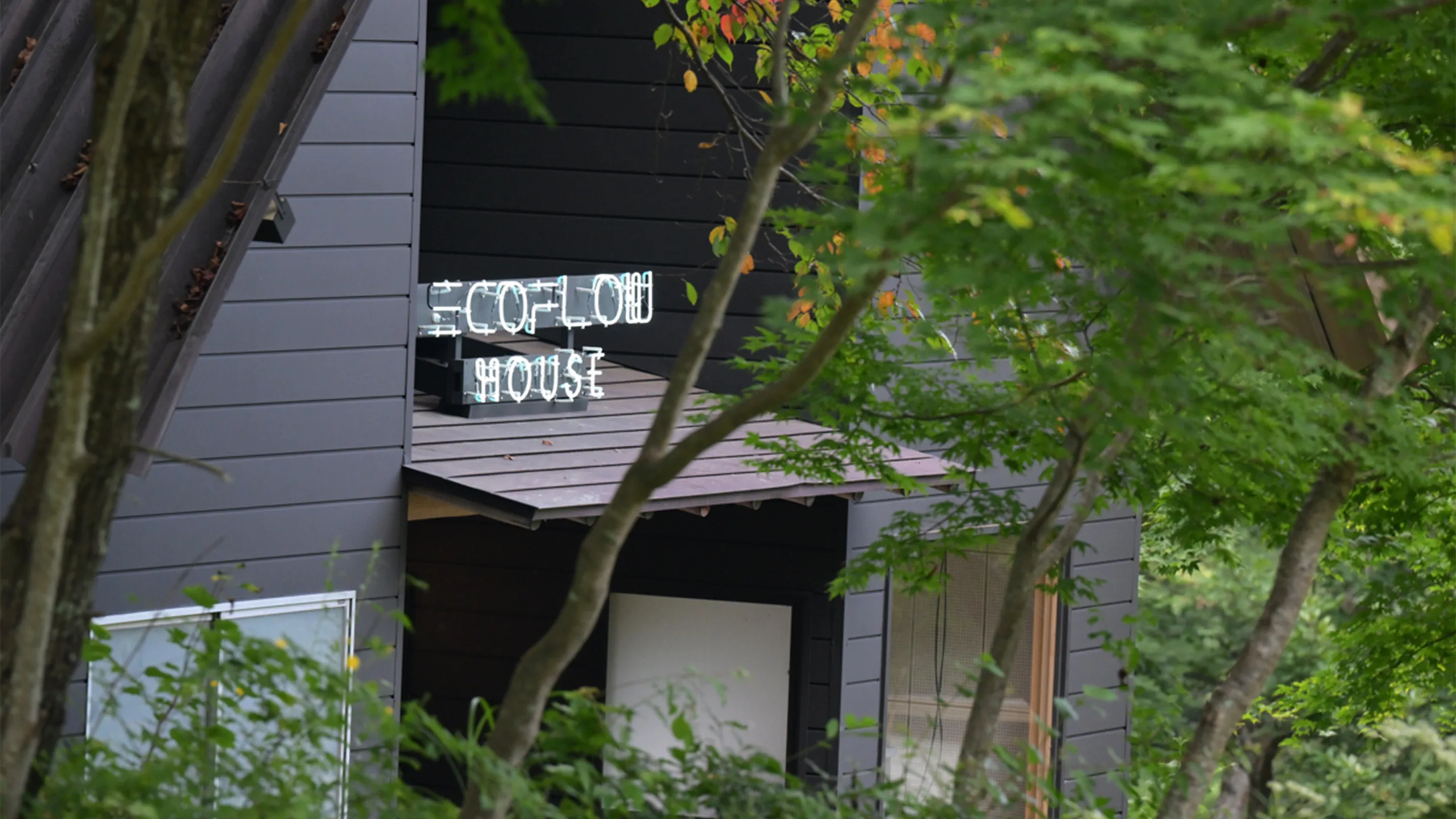 A dark-colored building is partially obscured by green trees. A neon sign on the porch roof reads ECOFLOW HOUSE. The setting appears natural and peaceful.