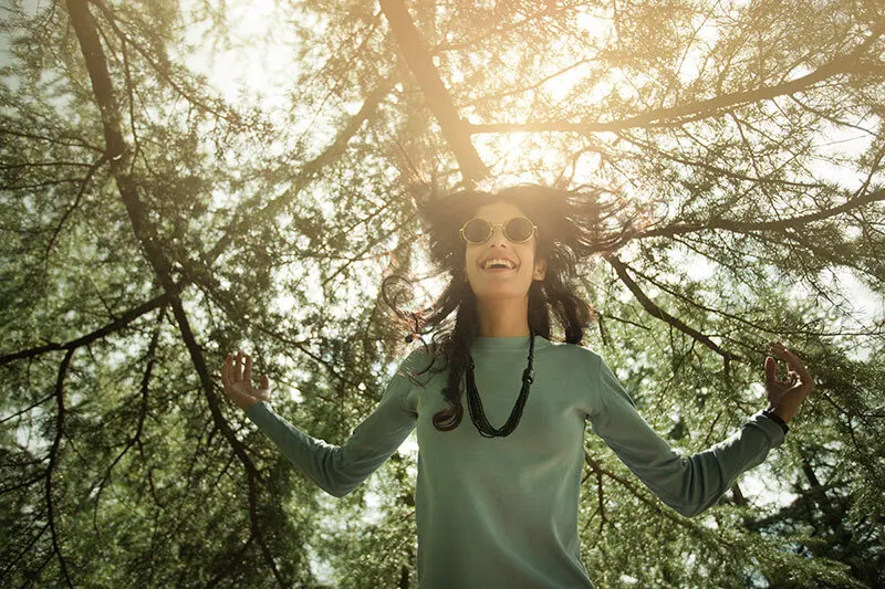 A woman wearing sunglasses and a long-sleeve shirt stands outdoors under sunlit tree branches, smiling with her arms raised, as sunlight filters through the leaves above her.