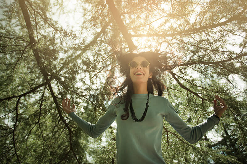 A woman wearing sunglasses and a long-sleeve shirt stands outdoors under sunlit tree branches, smiling with her arms raised, as sunlight filters through the leaves above her.