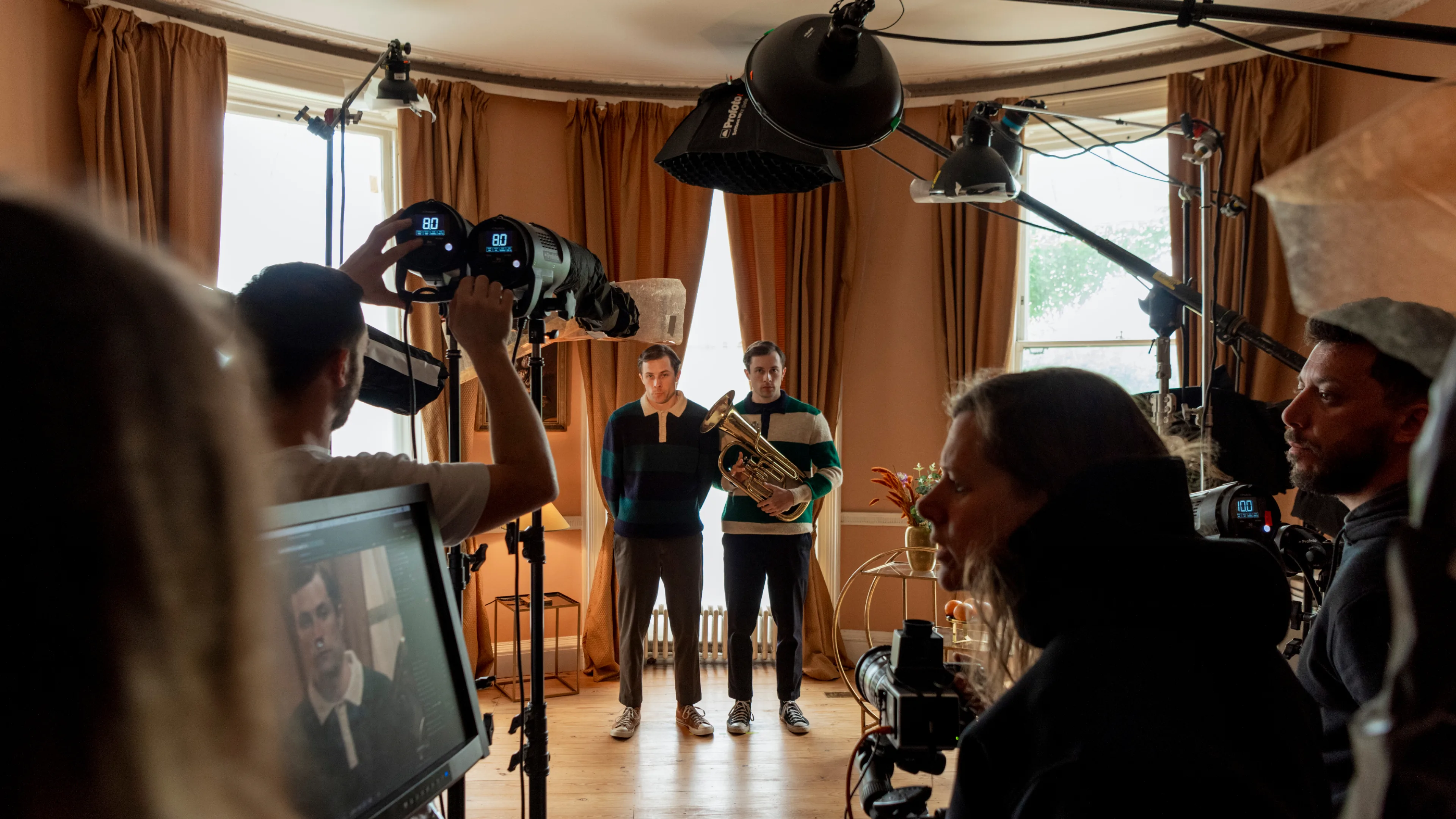 A film crew records two men standing close together in a warmly-lit room, one holding a French horn, surrounded by cameras, lights, and equipment. A monitor displays a close-up of one of the men.