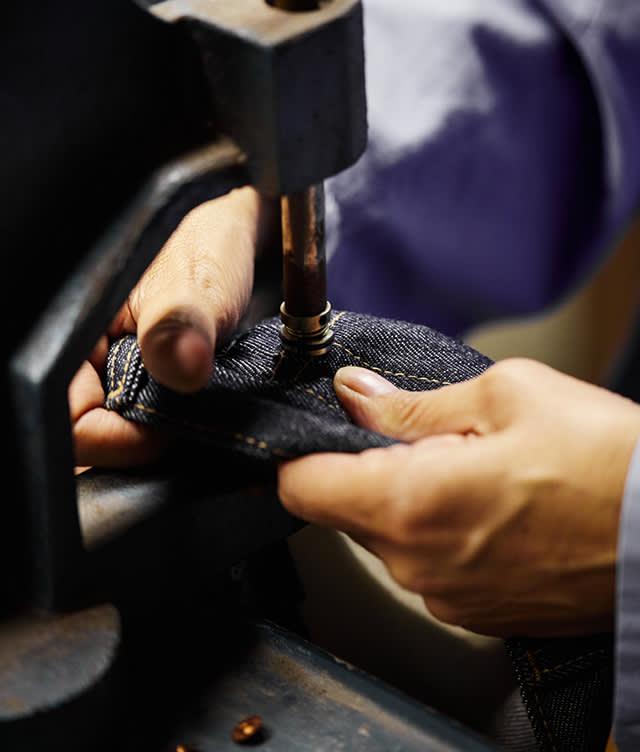 Close-up of hands using a machine to press a metal rivet into dark denim fabric, likely making or repairing jeans. The person is wearing a light blue shirt.