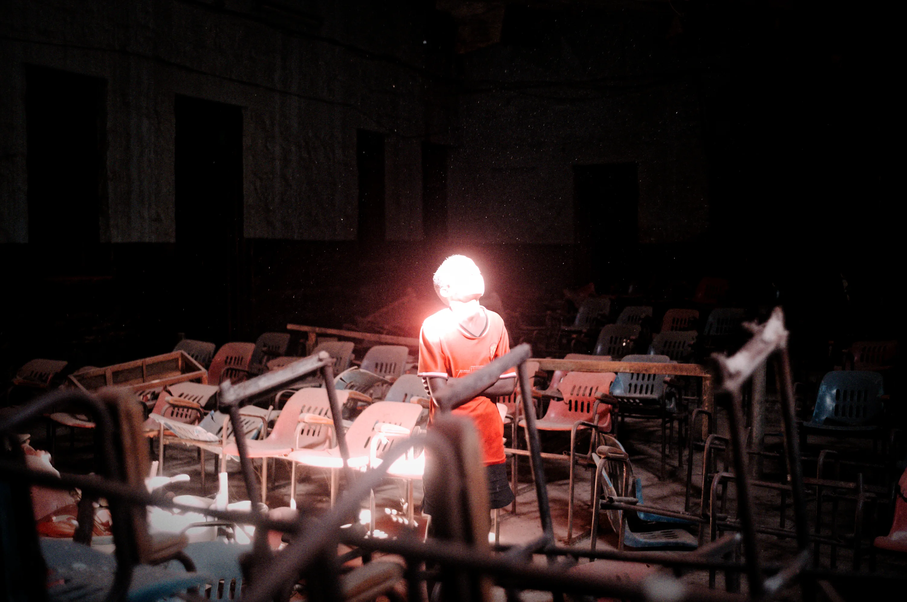 A person in a red shirt stands facing away, illuminated by a bright light, in a dark room filled with empty, disordered chairs and desks.