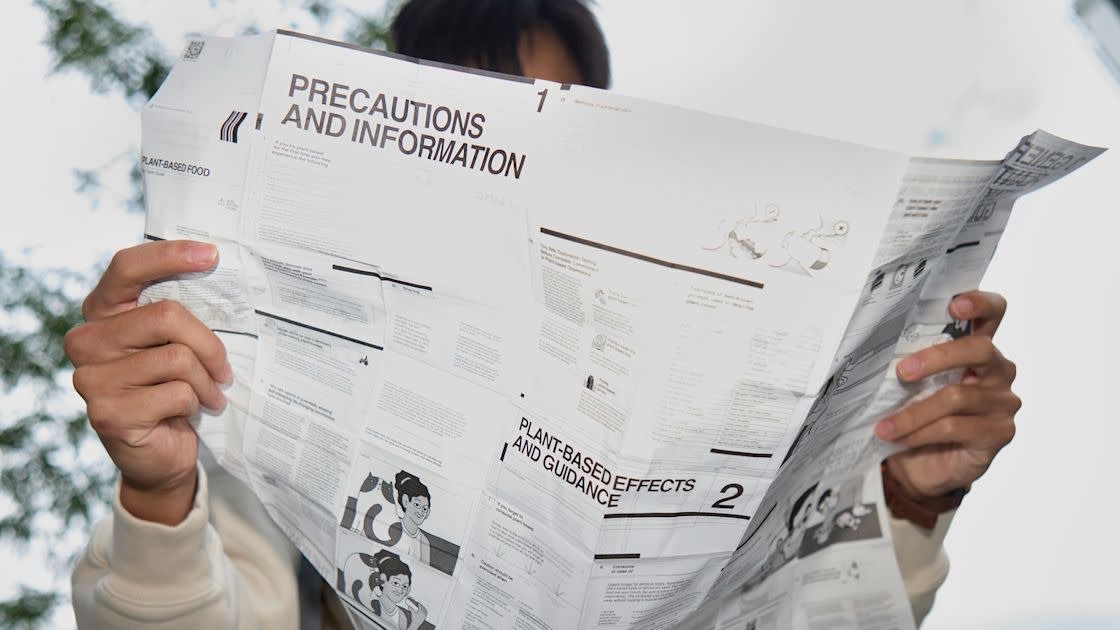 A person holds and reads a large, unfolded instruction leaflet labeled Precautions and Information outdoors, with only their hands and part of their face visible behind the paper.