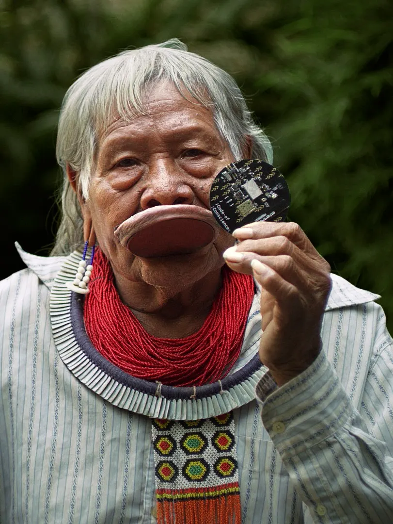 An elderly Indigenous person with long gray hair, wearing traditional jewelry and a lip plate, holds up a circular object resembling an electronic circuit board, standing against a blurred green background.