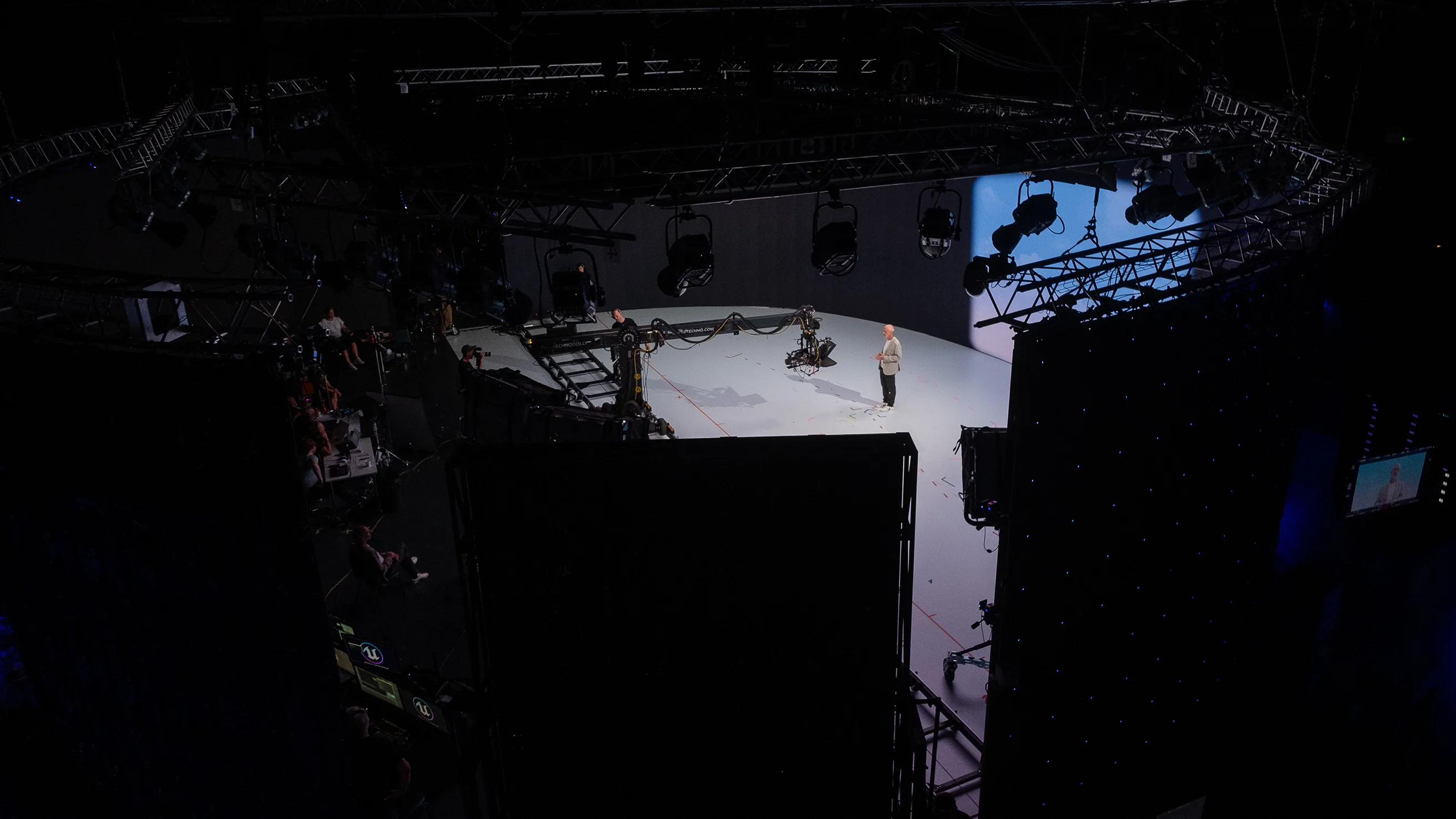 A person stands on a brightly lit stage in a large studio, surrounded by cameras, lighting equipment, and crew members, with a screen displaying their image in the background.
